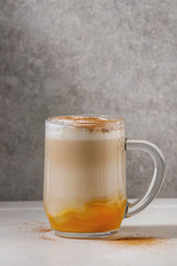 Glass of pumpkin layered spice latte with pumpkin puree, milk foam and cinnamon standing on white marble table with grey wall at background. Copy space