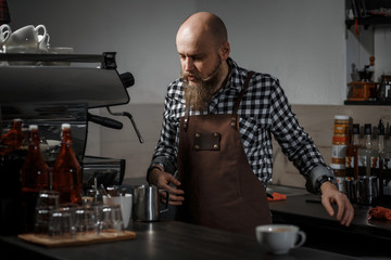 Brutal young barista in an apron makes coffee at the bar in a modern cafe