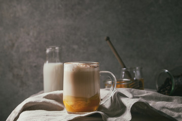 Glass of pumpkin layered spice latte with pumpkin puree, milk foam and cinnamon standing with ingredients in jars and decorative white pumpkins on crumpled table cloth with grey wall at background.