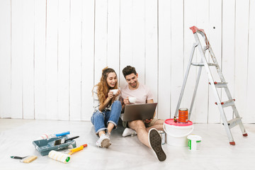 Photo of smiling couple man and woman sitting on floor over white wall near ladder and using...