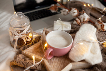 Mug of hot cappuccino on a wooden tray is on the bed. Cozy decor. Breakfast. Mug, plaid, cotton, laptop, candle. Gift box and knitted mittens. Christmas lights. Holidays. Christmas. Autumn. Winter.