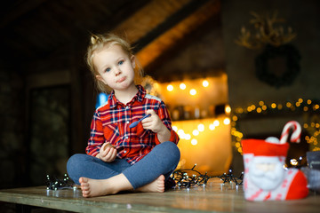 Fototapeta na wymiar Little white blonde girl sitting on a wooden table in the Chalet, decorated for Christmas tree and garlands with lights, and eating a chocolate egg from her christmas gift. Christmas morning concept.