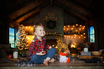 Fototapeta na wymiar A cute baby smiles and opens his sweet Christmas gift, sitting on a wooden table, in a decorated hall with a Christmas tree and garlands.