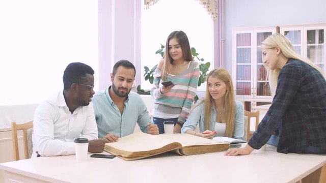 Handsome Arabian young bearded man surrounded by multiracial male and female students holding in hands big ancient book in library