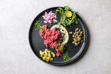Beef tartare with quail egg in shell, cutting pickled cucumbers, capers, red onion, chives, arugula salad served in black ceramic plate over grey background. Flat lay, space