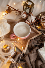 Mug of hot cappuccino on a wooden tray is on the bed. Cozy decor. Breakfast. Mug, plaid, cotton, candle. Gift box and knitted mittens. Christmas lights. Holidays. Christmas. Autumn. Winter.