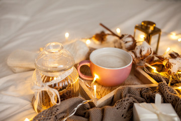 Obraz na płótnie Canvas Mug of hot cappuccino on a wooden tray is on the bed. Cozy decor. Breakfast. Mug, plaid, cotton, candle. Gift box and knitted mittens. Christmas lights. Holidays. Christmas. Autumn. Winter.