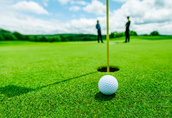 balle de golf dans l& 39 herbe verte près du trou avec l& 39 ombre du mât et deux personnes floues en arrière-plan