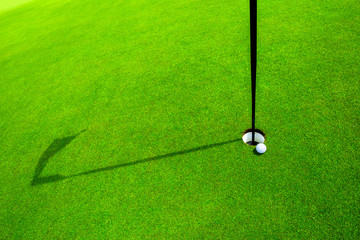 golf ball in the green grass close to the hole with the shadow of the flagpole