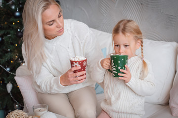 Young beautiful blonde woman hugs her little daughter on the couch with mugs of warm drink. Mother care about the child at home. The tenderness of my mother's embraces. Christmas winter comfort