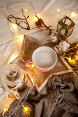 Mug of hot cappuccino on a wooden tray is on the bed. Cozy decor. Breakfast. Mug, plaid, cotton, candle. Book. Christmas lights. Holidays. Christmas. Autumn. Winter.