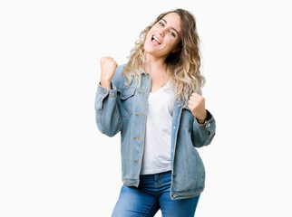 Beautiful young blonde woman wearing denim jacket over isolated background very happy and excited doing winner gesture with arms raised, smiling and screaming for success. Celebration concept.