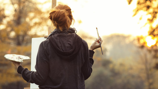 red-haired girl with painting a picture on an easel in nature, a young woman involved in creativity and enjoying beautiful landscape at sunset