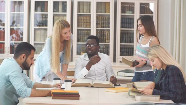 Pensive african american young man in stylish eyeglasses for vision correction, Arab man and three euopean young female students reading big old textbook sitting at the table against window .