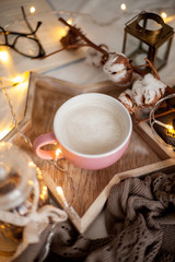 Mug of hot cappuccino on a wooden tray is on the bed. Cozy decor. Breakfast. Mug, plaid, cotton, candle. Book. Christmas lights. Holidays. Christmas. Autumn. Winter.