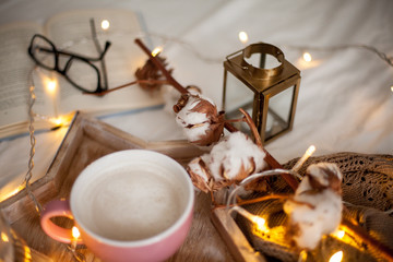 Fototapeta na wymiar Mug of hot cappuccino on a wooden tray is on the bed. Cozy decor. Breakfast. Mug, plaid, cotton, candle. Book. Christmas lights. Holidays. Christmas. Autumn. Winter.