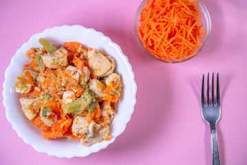 stewed chicken, with vegetables, carrots, cabbage on a plate. next to grate fresh carrots. pink background.