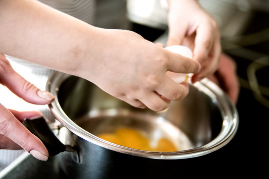 A child helps mom cook breakfast and breaks eggs into a saucepan.
