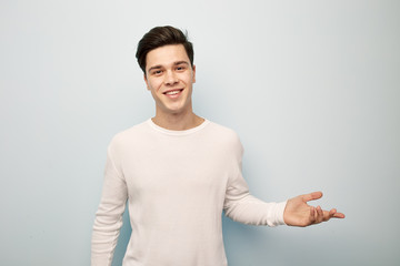 Smiling dark-haired guy dressed in a white long sleeve t-shirt points his hand to the side on a white background