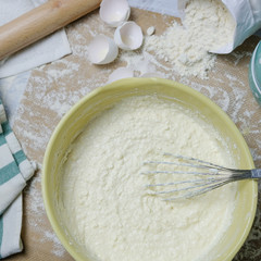 Table with ingredients for dough, raw eggs, flour, plate, napkins, rolling pin and whisk. Process of mixing, preparing pie, kitchen objects, square flat lay with light colors.