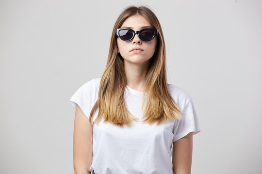 Serious girl dressed in a white t-shirt and sun glasses is on a white background in the studio
