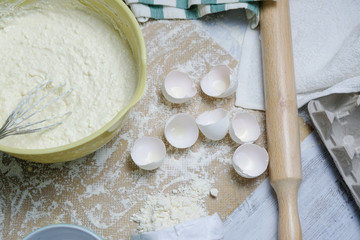 Table with ingredients for dough, raw eggs, flour, plate, napkins, rolling pin and whisk. Process of mixing, preparing pie, kitchen objects, flat lay with light colors.