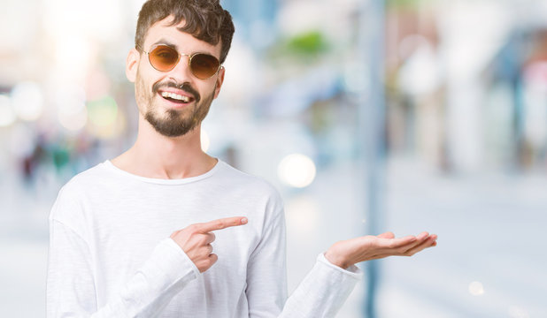 Young handsome man wearing sunglasses over isolated background amazed and smiling to the camera while presenting with hand and pointing with finger.