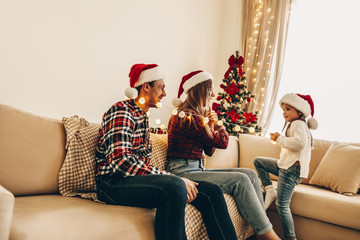 Christmas. Family. Happiness. Little girl and her parents in Santa hats are having fun while sitting at home near the Christmas tree