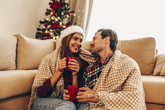 Christmas. Love. Home. Young couple in Santa hats, covered in plaid is holding cups, talking and smiling while sitting at home near the Christmas tree