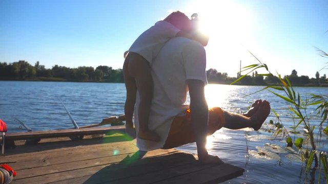 Young man practicing yoga exercise with son on his back on the edge of wooden jetty at lake. Family spending time together at nature on summer day. Healthy active lifestyle. Slow motion Close up