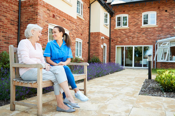 Senior Woman Sitting On Bench And Talking With Nurse In Retirement Home - obrazy, fototapety, plakaty