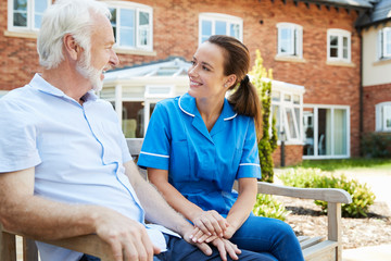 Senior Man Sitting On Bench And Talking With Nurse In Retirement Home