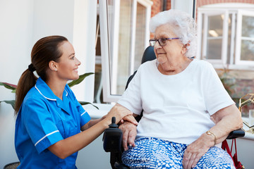Senior Woman Sitting In Motorized Wheelchair Talking With Nurse In Retirement Home