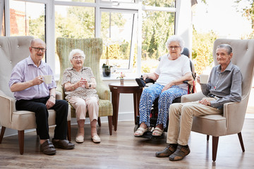Portrait Of Male And Female Residents Sitting In Chairs In Lounge Of Retirement Home