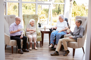 Male And Female Residents Sitting In Chairs And Talking In Lounge Of Retirement Home