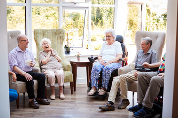 Male And Female Residents Sitting In Chairs And Talking In Lounge Of Retirement Home - obrazy, fototapety, plakaty
