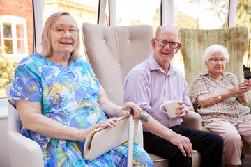 Portrait Of Male And Female Residents Sitting In Chairs In Lounge Of Retirement Home