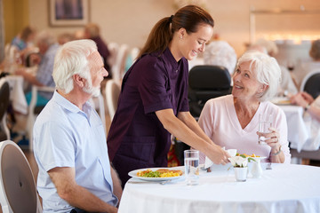 Senior Couple Being Served With Meal By Carer In Dining Room Of Retirement Home - Powered by Adobe