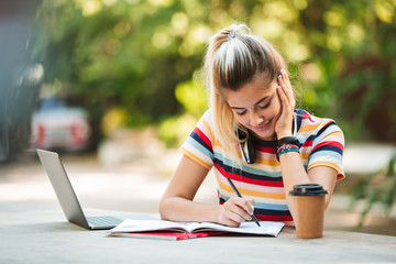 Happy young cute girl student sitting in park using laptop computer.