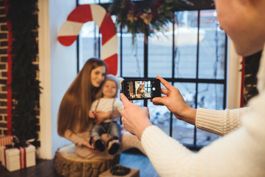 Theme mobile photography, amateur photo and video on phone. Hands Caucasian man holds uses smartphone makes photo of mother and son at home near window in winter in Christmas time New Years decor