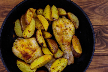 Baked chicken drumsticks with potatoes in in cast-iron frying pan on wooden table. Top view