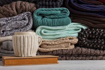 A stack of knitted clothes of different colors and textures, on the table next to a book and a cup in a case and a place for text. Copy space.