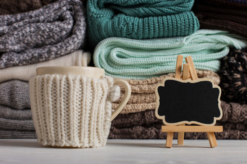 A stack of knitted clothes of different colors and textures, on the table next to a cup in a case...