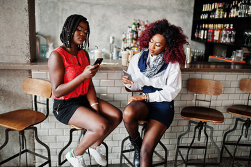 Attractive african american two girls friends sitting at chairs against bar on pub and looking at mobile phones.