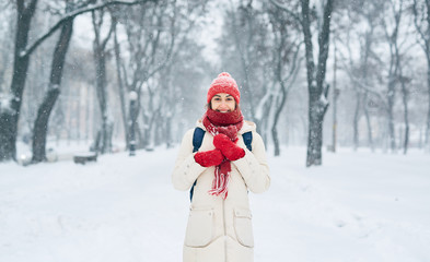 Fototapeta na wymiar portrait of Cheerful smiling woman in white down jacket and red cap, scarf and mittens on the snowy alley after blizzard in city. copyspace