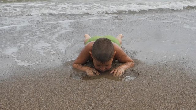 child lies on his stomach on the sandy shore in the sea waves. Boy rowing sand on the beach. Kid washed sea waves.