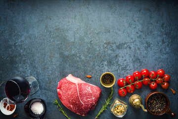 Raw marbled beef steak on dark concrete background with salt and herbs, tomatoes and glass of red wine. Top view and copyspace