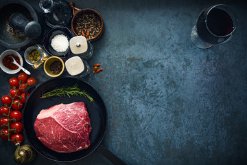 Raw marbled beef steak on cast iron pan with salt and herbs, tomatoes and glass of red wine. Top view and copyspace.