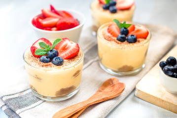 homemade, exquisite dessert tiramisu in glasses decorated with strawberry, blueberry, mint on white wooden table.