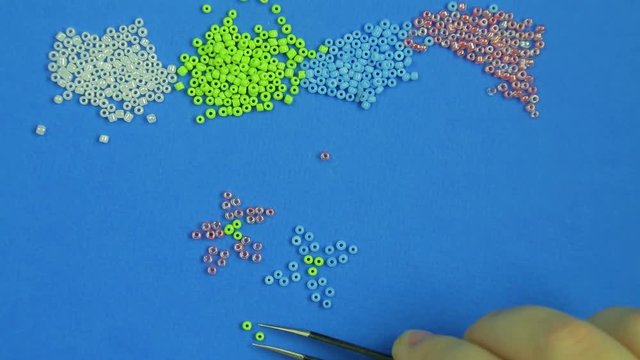 Female hand on a blue background pokes out flowers from multi-colored beads with tweezers. Close-up